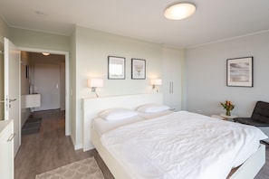 Una habitación de hotel moderna con una cama grande, mesitas de noche, un escritorio y una silla. Hay cuadros en la pared y una pequeña mesa con un jarrón de flores.