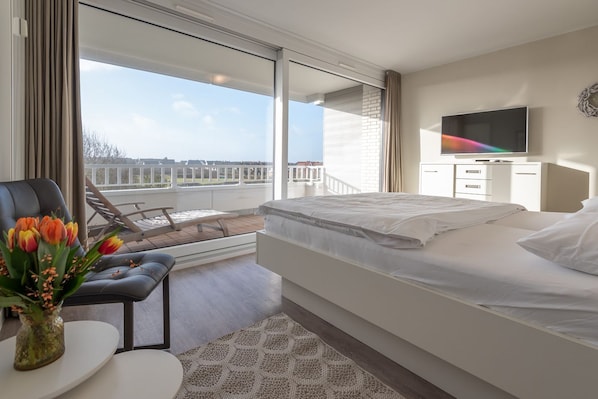 A hotel room with a bed, a TV, a desk, and a balcony with a view. - Lichtblick im Haus Cäcilia - Modern 2-room apartment with beautiful balcony (Wenningstedt-Braderup (Sylt))