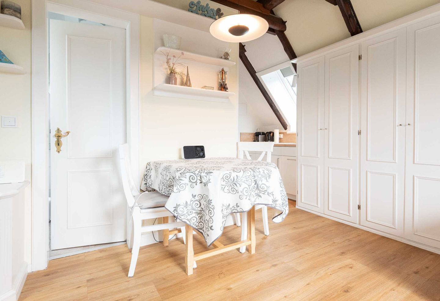 Un coin repas avec une table dressée pour deux personnes, des chaises blanches, une nappe à motifs, un luminaire au plafond et des meubles blancs.