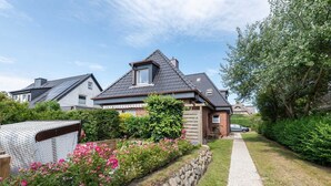 A residential house with a sloped roof, a visible attic window, and a garden with pink flowers and a stone wall. - Sylt feel-good nest, App. 2 - Cozy 1-room apartment with approx. 25 m², (Wenningstedt-Braderup (Sylt))