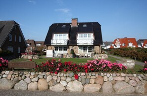 A large, two-story house with a prominent balcony, surrounded by a well-maintained garden with pink flowers and a stone wall.