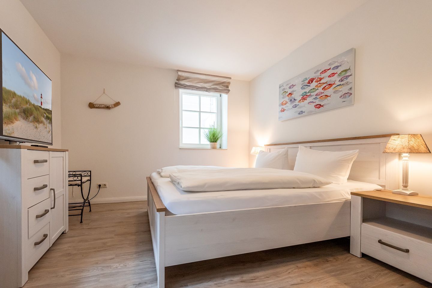 Un dormitorio con cama, mesita de noche, lámpara, televisor, cómoda, ventana y obra de arte.
