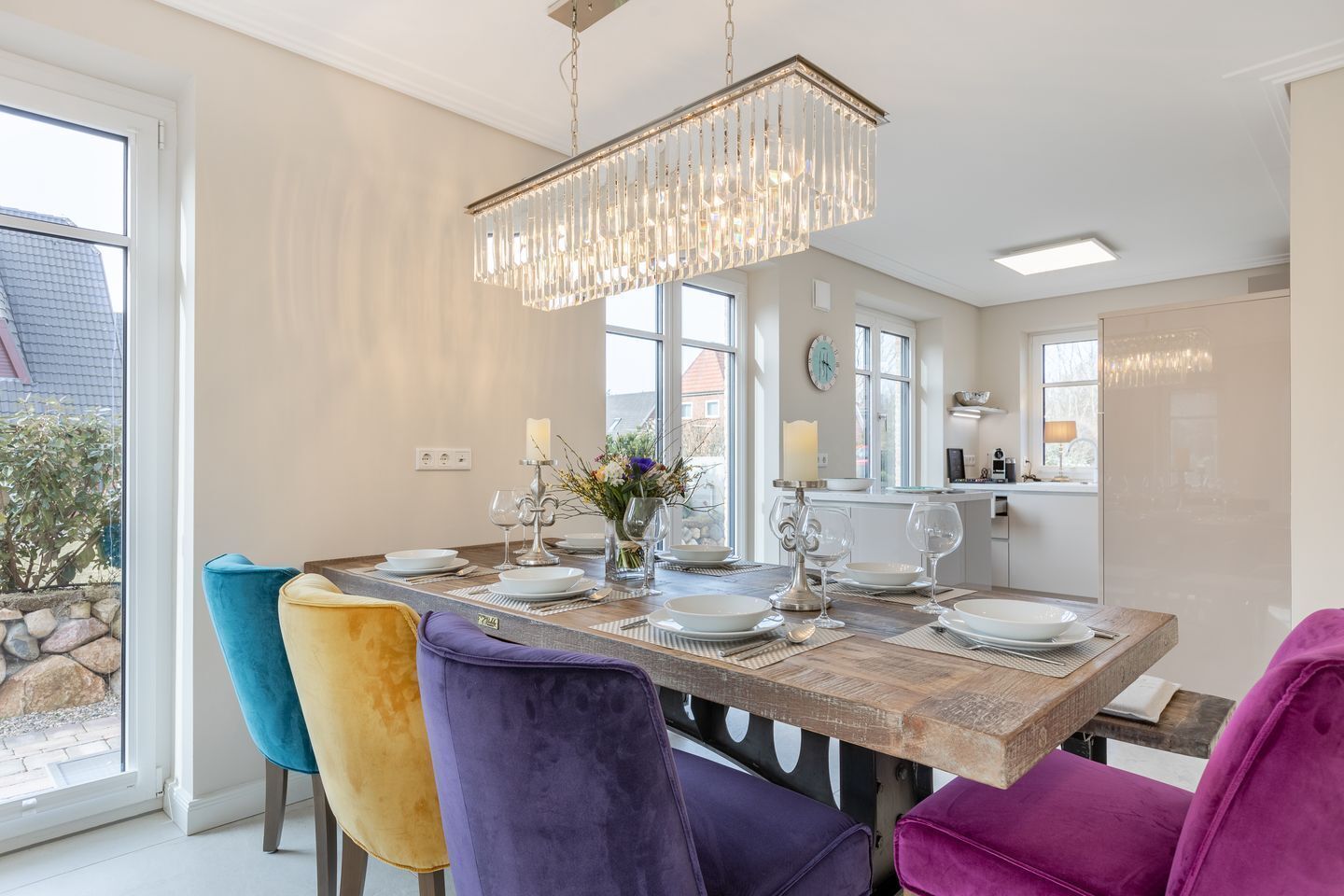Un coin repas avec une table en bois dressée pour quatre personnes, entourée de chaises aux couleurs vives, d’un lustre et offrant une vue sur l’extérieur.