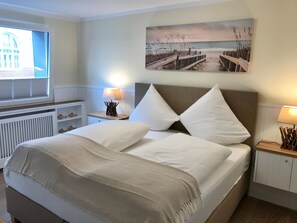 Ein aufgeräumtes Schlafzimmer mit einem großen Bett, einem Fenster mit Jalousien, einem Strandbild und Nachttischlampen.