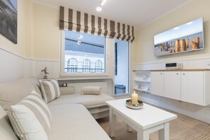 En hyggelig stue med sofa, et bord med et stearinlys, et vindue med udsigt til bygninger og et fjernsyn på væggen.