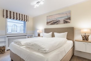 En pænt redt seng med hvide sengetæpper, en sengegavl, sengeborde med lamper, et vindue med gardiner, en radiator og et billede af en strand.
