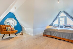 A bedroom with a sloped ceiling, a bed with a grey and orange bedspread, a blue accent wall, and a round window.