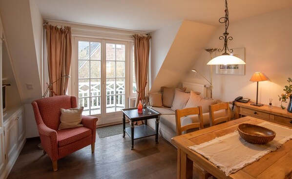 Una sala acogedora con un sillón rojo estampado, una mesa de comedor de madera con un tazón, una mesita auxiliar y una ventana con cortinas.