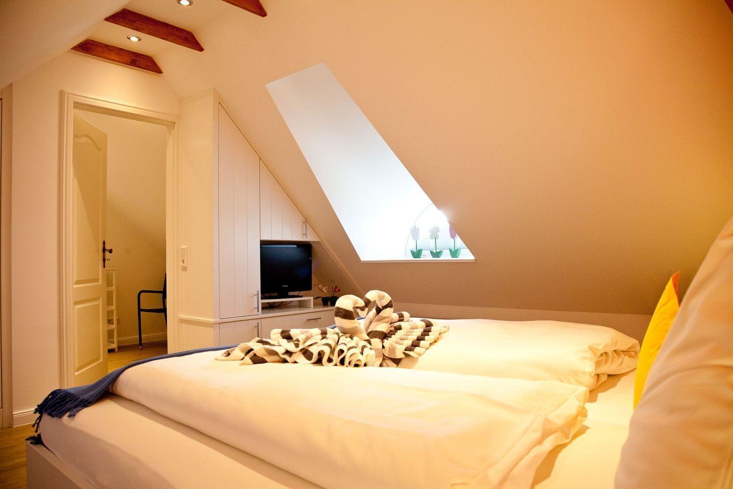Een slaapkamer met schuine wand, een bed met een gestreept dekbed, een televisie op een standaard en een raam met uitzicht op bloemen.