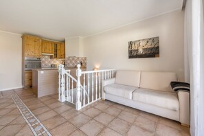 Una sala de estar con sofá, una escalera con barandilla blanca, una cocina con armarios de madera y piso de baldosas.