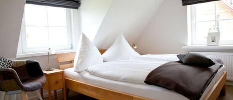 A bedroom with a wooden bed, white bedding, a desk with a chair, and large windows with dark curtains.