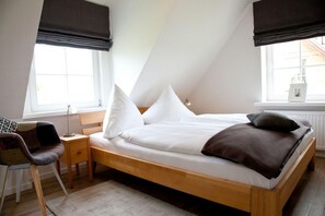 Quarto com cama de madeira, roupa de cama branca, escrivaninha com cadeira e janelas amplas com cortinas escuras.