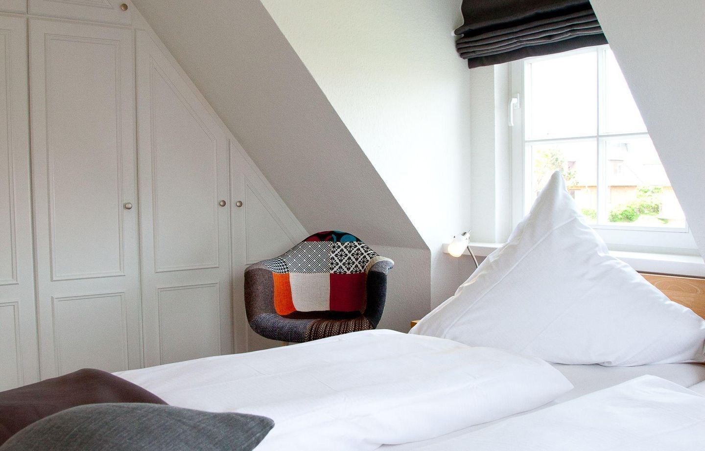 A bedroom with a sloped ceiling, a bed with white bedding, a colorful ottoman, and a window with a view of greenery.