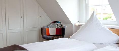 A bedroom with a sloped ceiling, a bed with white bedding, a colorful ottoman, and a window with a view of greenery.