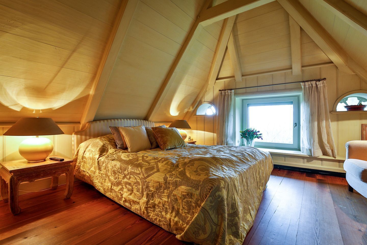Quarto com teto inclinado, cama com colcha estampada, mesa de cabeceira com abajur, janela com cortinas e vista para o exterior.