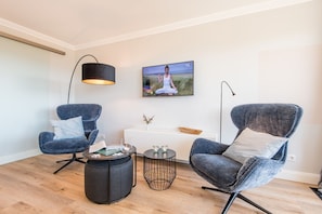 Un salon moderne avec deux fauteuils bleus, une petite table basse ronde, une table d’appoint, un lampadaire, un tableau et une console blanche.
