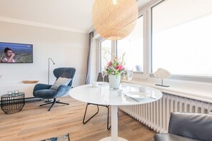 Un salon moderne avec une table ronde blanche, un fauteuil bleu, un lampadaire et une grande fenêtre donnant sur un beau paysage.