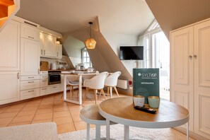 Une cuisine moderne avec des meubles blancs, un coin repas avec des chaises blanches, une table avec un service à café et un téléviseur fixé au mur.