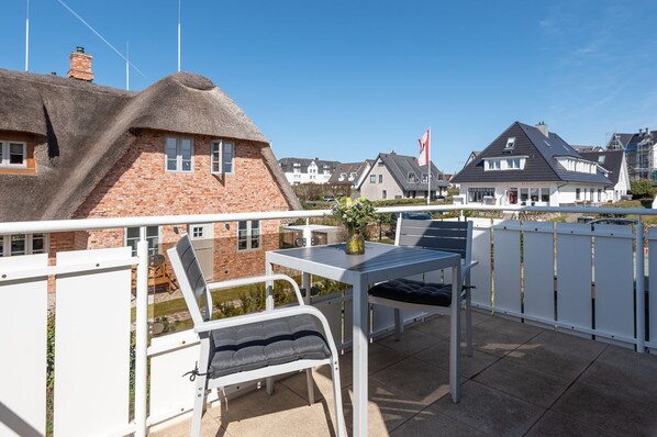 Un balcon avec une table et des chaises, une maison à toit de chaume en arrière-plan et un drapeau sur un mât.