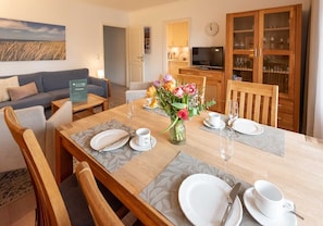 A dining area with a wooden table set for four, a vase of flowers, and a television in the background. - Haus Atrium Apartment 19 - Cozy 2-room vacation apartment with approx. 45 m², for (Sylt)