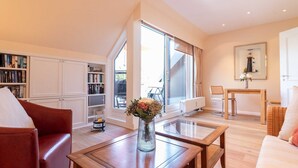 A spacious living room with a large window, a bookshelf, a coffee table, and a sofa. - Käpt`n Nelson, App 10 - Bright 2-room vacation apartment with approx. 52 m² living space, (Wenningstedt-Braderup (Sylt))