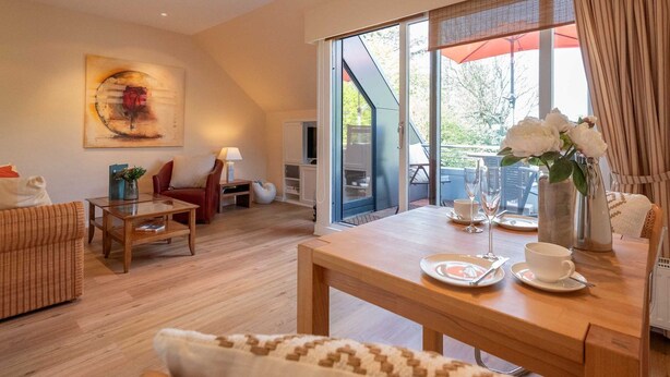 A modern living room with a wooden floor, a sofa, a coffee table, and a dining table set with a vase of flowers and a glass of wine. - Käpt`n Nelson, App 10 - Bright 2-room vacation apartment with approx. 52 m² living space, (Wenningstedt-Braderup (Sylt))