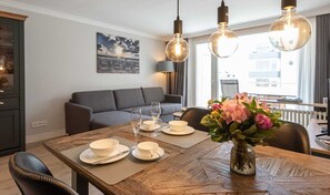 A dining area with a wooden table set for four, a vase of flowers, and a modern sofa in the background. - Apartment Eydum Nr. 38 - Beautiful 2-room apartment in city location on approx. (Sylt)