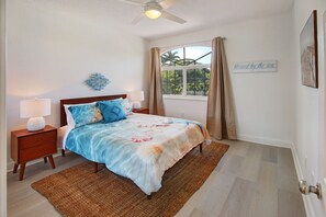 Une chambre à coucher comprenant un lit, des tables de chevet, une fenêtre avec des rideaux, un ventilateur de plafond et un panneau décoratif mural.