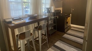 Eine kleine Küche mit dunkler Arbeitsplatte, einem Gasherd, einem Spülbecken und einem Fenster mit Vorhängen. Zwei Hocker und ein kleiner Tisch mit einer Schüssel sind ebenfalls vorhanden.