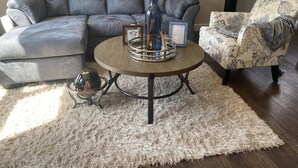 Ein Wohnzimmer mit einem grauen Ecksofa, einem runden Couchtisch mit Ablage, einem gemusterten Sessel und einem flauschigen Teppich.