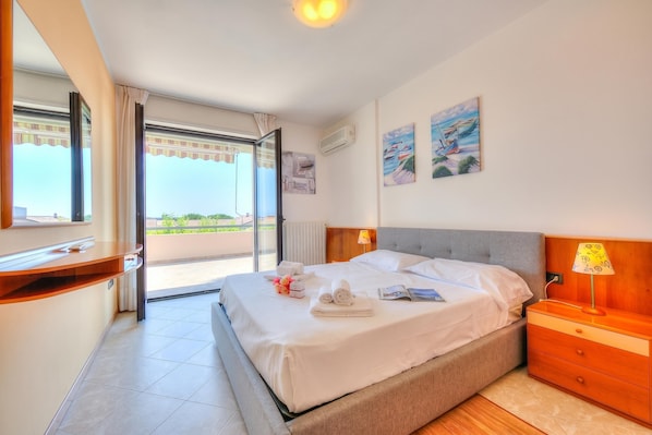 A hotel room with a bed, bedside table, lamp, artwork, and a view through a window.