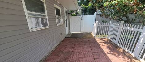A covered patio with red brick flooring, a white fence, and a house with grey siding and white trim.