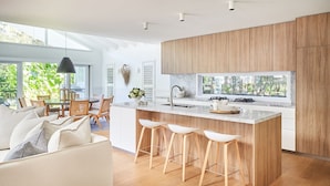 A modern kitchen with a large island, wooden cabinets, and a view of greenery outside. - This home matches quaint beachside charm with luxurious comfort. (Palm Beach)