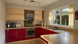 A modern kitchen with wooden cabinets, a red countertop, stainless steel appliances, and a window with a view of greenery. - 2 River Court, Invergarry (Invergarry)