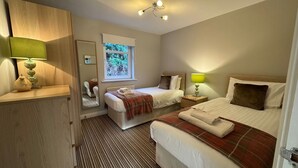 A hotel room with two beds, a wooden cabinet, a mirror, a window with a view of trees, and a striped carpet. - 2 River Court, Invergarry (Invergarry)