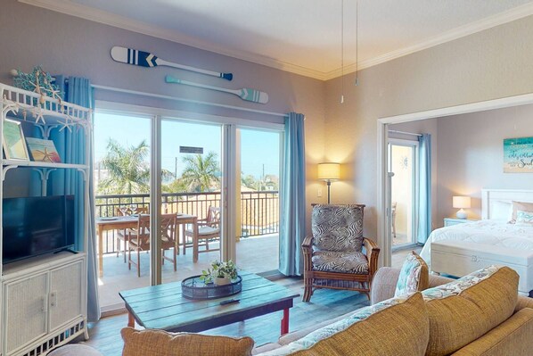 A hotel room with a balcony, a bed, a sofa, a chair, a TV, and a table with a plant. - Beach Access. Heated Pool and Hot Tub. Relaxation. Comfort. Views. Convenience. (Treasure Island)