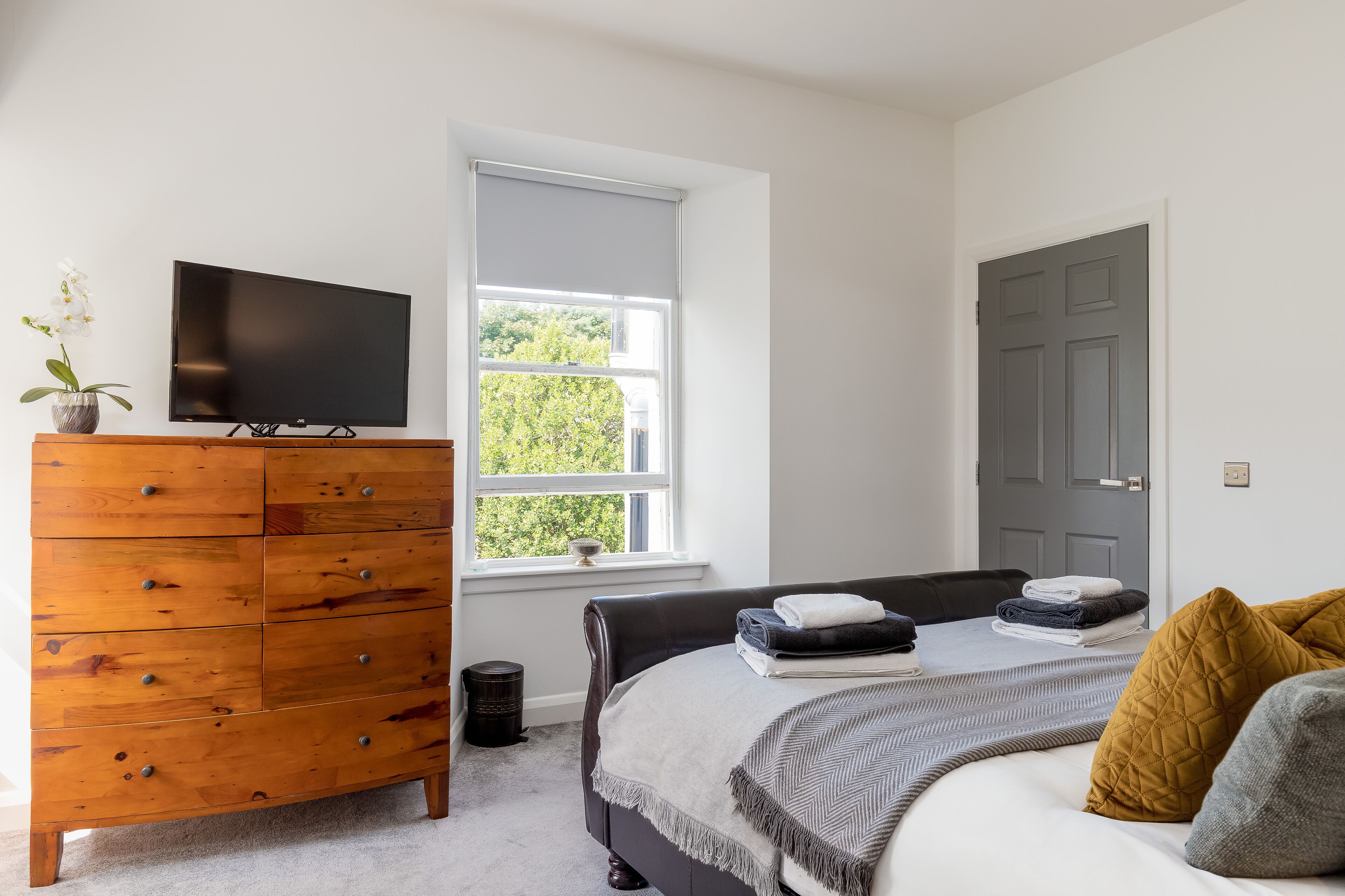 Um quarto com cama, cómoda de madeira, televisão, janela e porta.