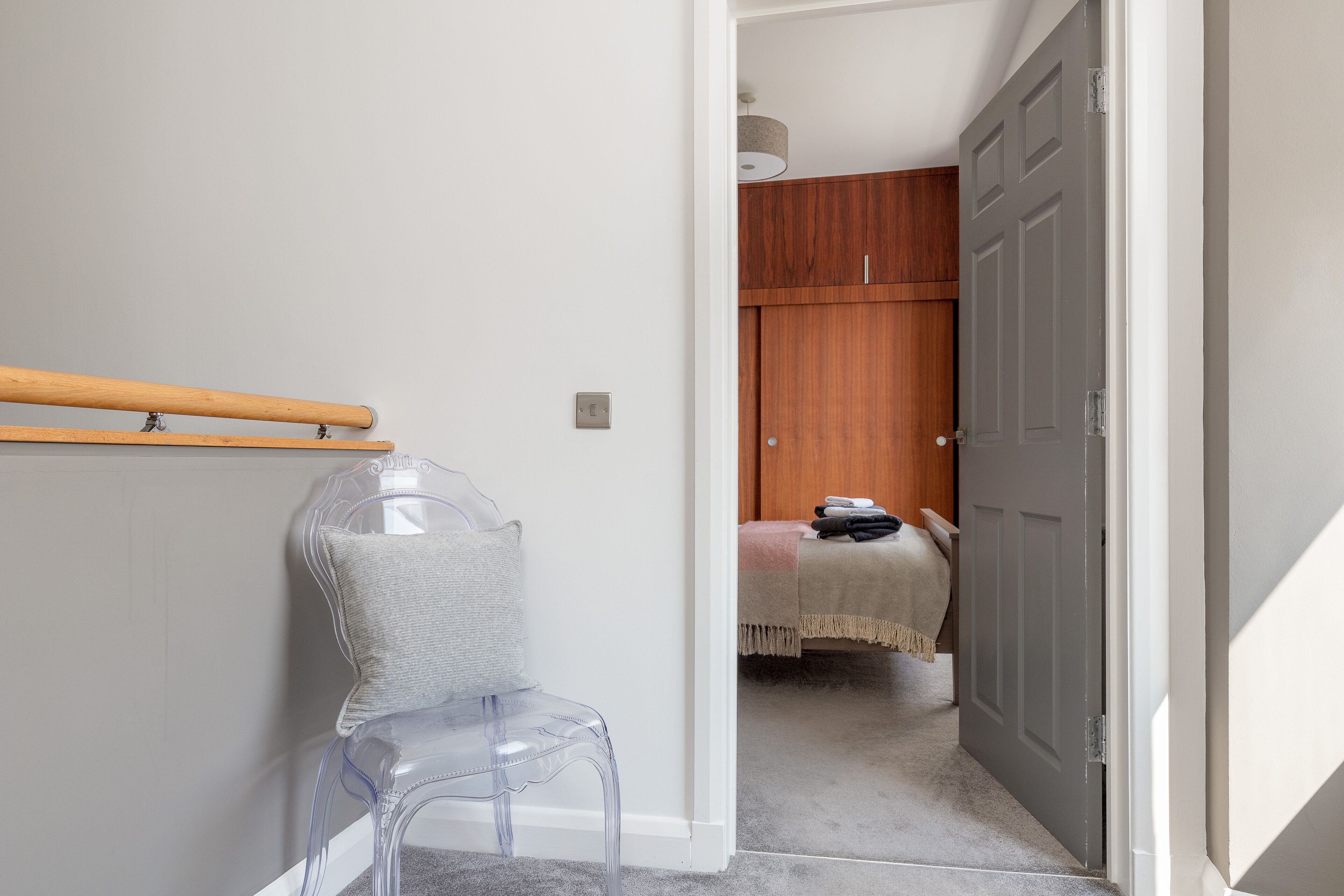 A room with a clear plastic chair, a wooden bench, and a grey door leading to another room with a bed and a wooden wardrobe.