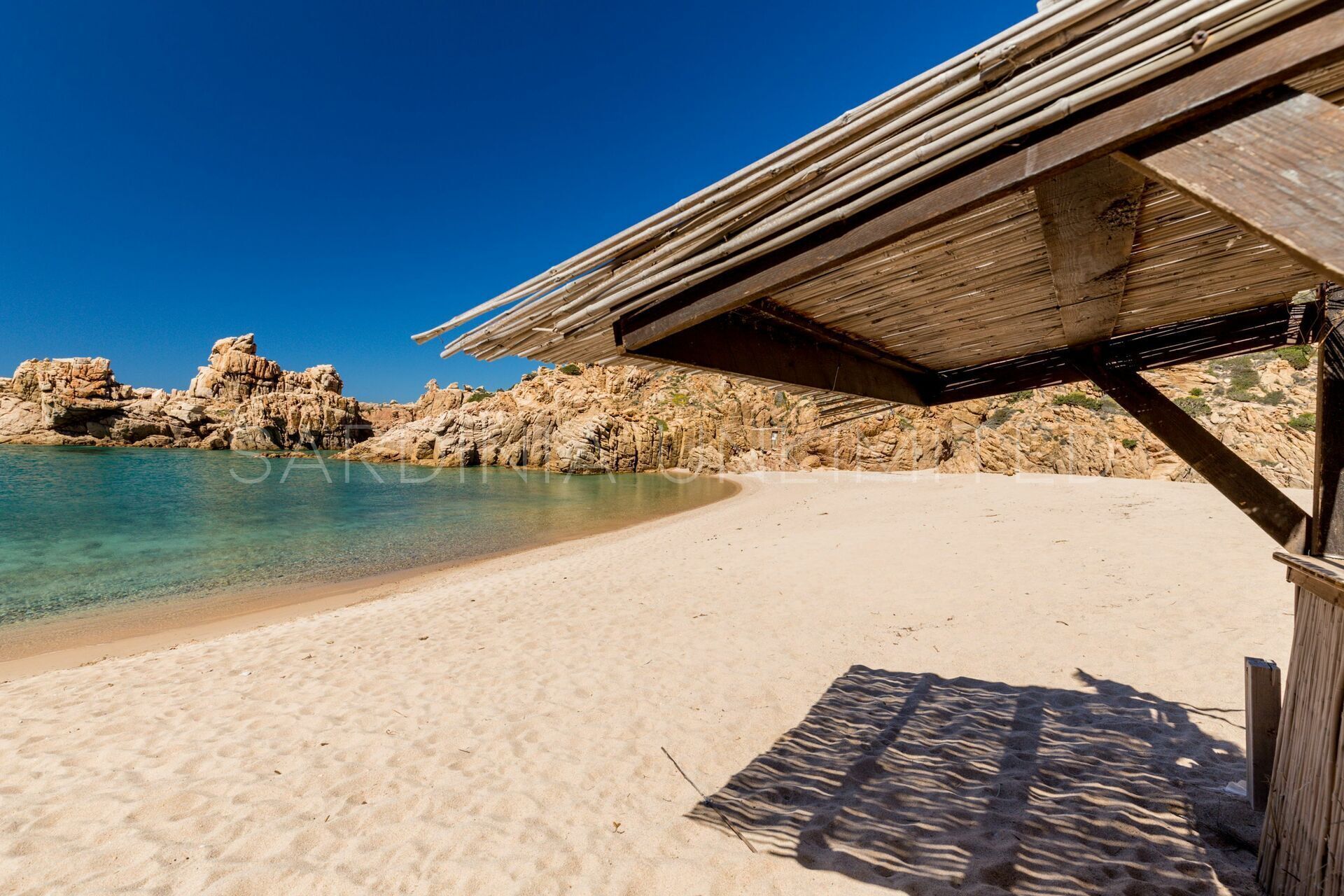 A sandy beach with clear water, rocky cliffs, and a wooden structure with a roof.