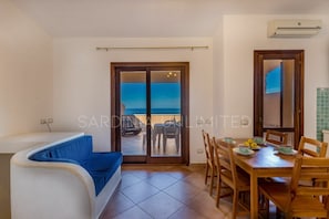 Una habitación con un sofá azul, una mesa de comedor de madera con sillas, vista al mar a través de un ventanal, y un equipo de aire acondicionado en la pared.