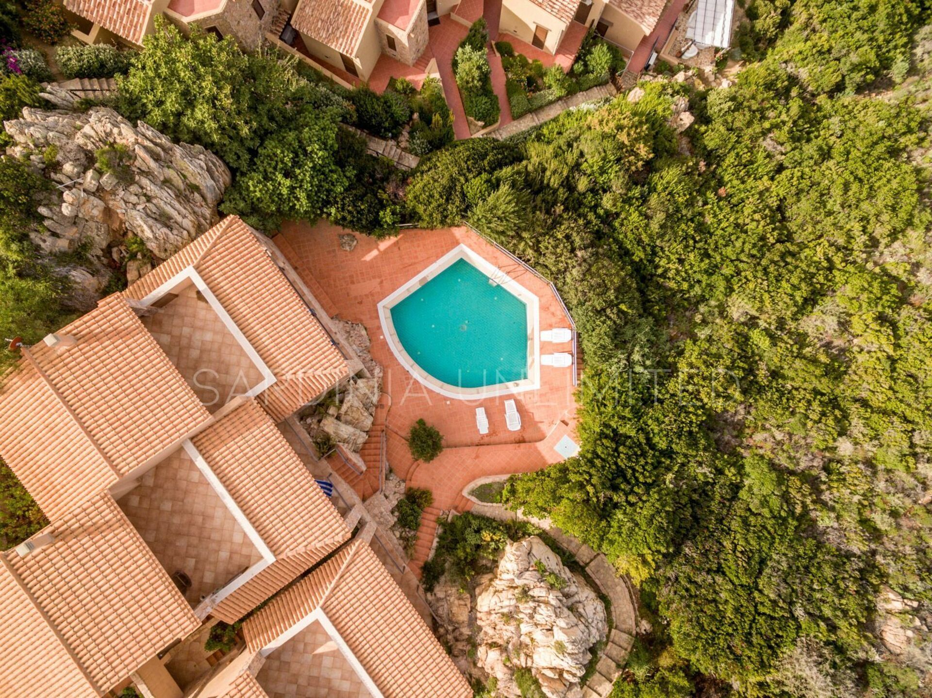 An aerial view of a resort with a swimming pool surrounded by lush greenery and terraced buildings with tiled roofs.