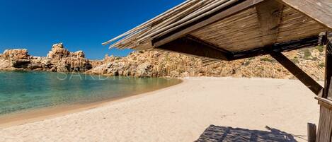 A sandy beach with clear water, rocky cliffs, and a wooden structure with a roof.