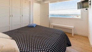 Un dormitorio con cama, un banco, un televisor y una ventana con vista al mar.