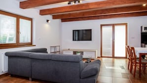 Una sala de estar moderna con un sofá seccional gris, vigas de madera en el techo, un televisor de pantalla plana y una zona de comedor con mesa y sillas.