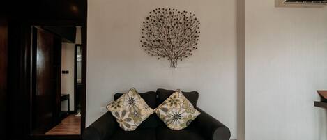 A modern living room with a dark sofa, two patterned throw pillows, and a wall-mounted abstract art piece.