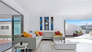Ein modernes Wohnzimmer mit einem grauen Ecksofa, einem weißen Couchtisch und einem Glastisch. An der Wand hängen drei abstrakte Gemälde und es bietet sich ein Blick ins Freie.