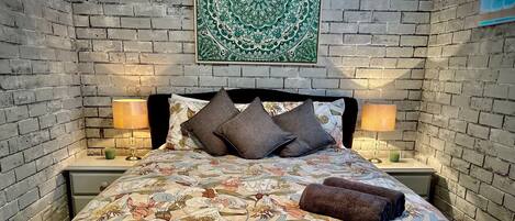 A bed with floral bedding, two bedside lamps, and a green patterned wall hanging.
