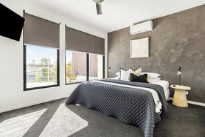 A modern bedroom with a large bed, a side table, a wall-mounted air conditioner, and a city view through large windows.