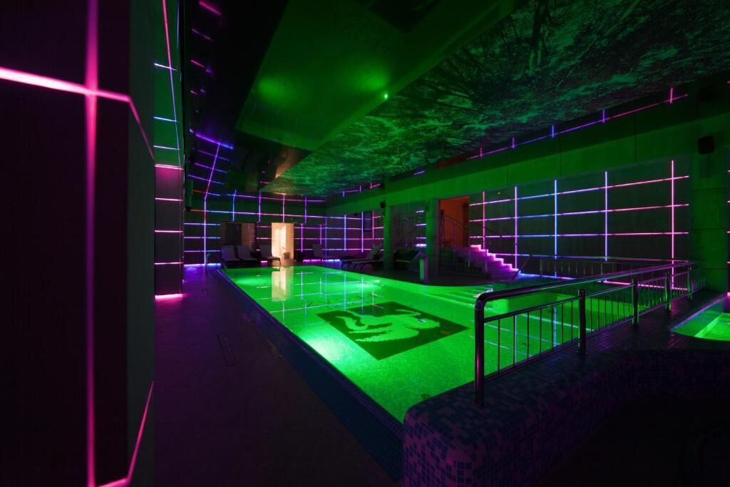Espaço interior moderno e com iluminação fraca, com área de piscina, iluminação neon e escadaria para um andar superior.
