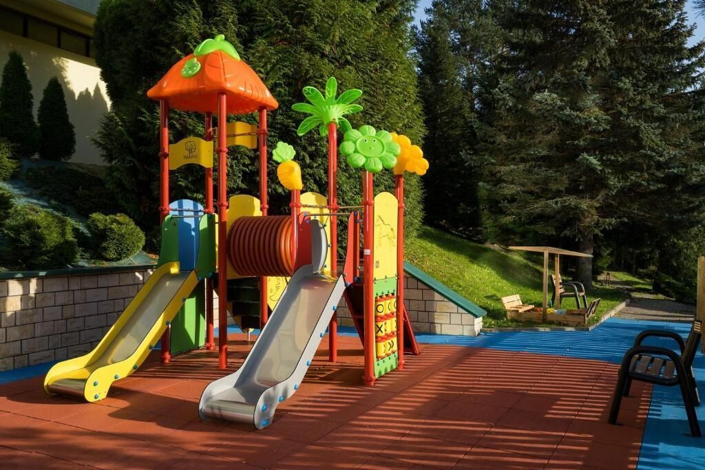 A playground with slides and climbing structures, surrounded by trees and a building in the background.